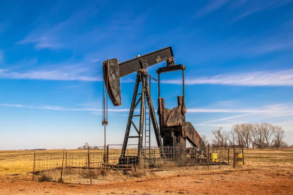 Black oil well pumpjack in brown field surrounded by metal fence.